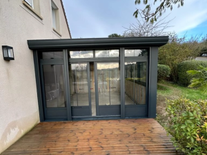 Extension anthracite vitrée sur terrasse bois, jardin palmier et ciel bleu