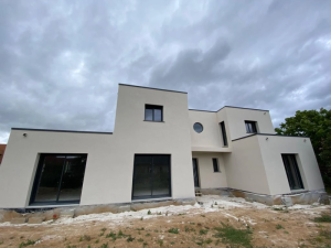 Maison cubique crepi beige, baies noires et fenetre oeil-de-boeuf sur terrain nu