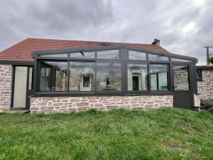 Véranda anthracite vitrée sur soubassement pierre, maison pierre et pelouse