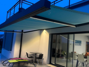 Store banne cyan eclaire par LED, facade blanche et baie vitree ouverte la nuit