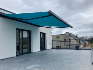 Store banne turquoise sur terrasse carrelée grise, garde-corps noir et toits voisins