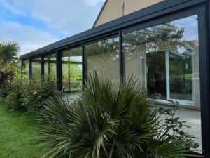 Gros plan veranda anthracite, reflets verdure dans vitrages et porte coulissante blanche