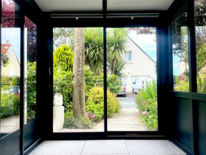Intérieur verrière anthracite carrelée, baie coulissante sur jardin et allée bois