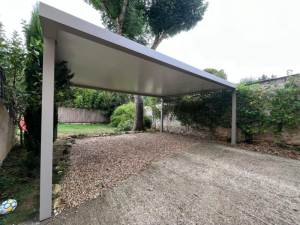 Carport aluminium gris clair toit plat sur allée béton, gravier et mur en pierre