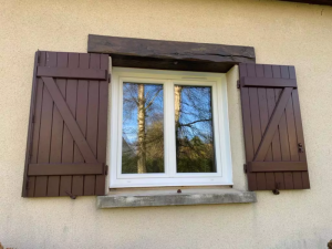 Fenêtre PVC blanche double vantail, volets bruns ouverts, crépi beige et linteau bois