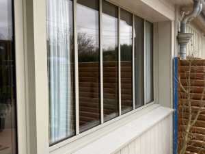 Fenetre quatre vantaux beige, reflet claustra bois et gouttiere sur bardage clair