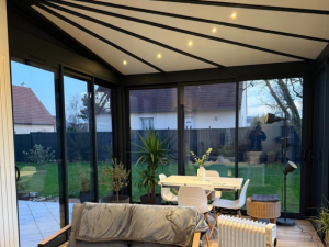 Véranda au crépuscule, baies anthracite, plafond poutres noires et spots chauds allumés