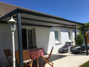 Pergola aluminium gris adossée, toile claire, lampe blanche et table bois sur terrasse