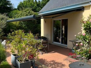 Store banne anthracite déployé sur terrasse pavée, table grise et rosiers roses
