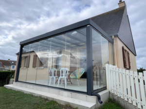 Véranda vitrée noire, table bois et chaises blanches devant maison à volets bruns