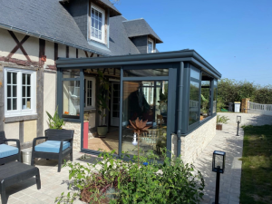 Véranda anthracite sur maison colombages, muret pierre et terrasse pavée avec salon gris
