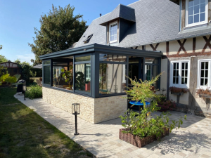 Véranda gris anthracite adossée aux colombages, muret pierre claire et terrasse pavée