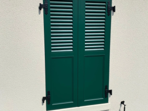 Volets battants verts mi-persiennés, pentures noires décoratives sur mur crépi clair