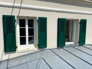 Deux fenêtres blanches, volets verts mi-persiennés sous bandeau blanc et toit zinc