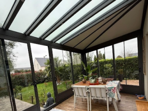 Interieur veranda anthracite, table bois, jardin haie palmiers et bouddha