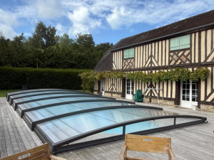 Abri bas cintré gris fermé sur piscine, colombages verts et haie dense
