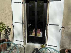 Porte-fenêtre cintrée noire, volets blancs ouverts et chaises turquoise sur terrasse pierre