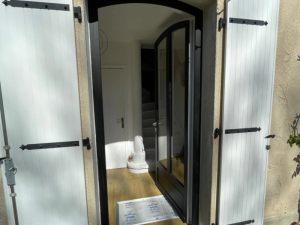 Porte cintrée noire entrouverte, volets blancs à pentures noires sur mur beige