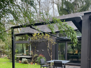 Armature pergola noire sur terrasse bois devant maison bardage sombre et jardin