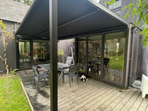 Store banne noir déployé sur terrasse bois, table grise et chien à l'ombre