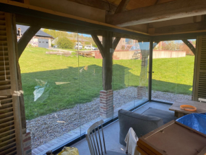 Rideau de verre sous préau bois, canapé gris, table et pelouse au fond