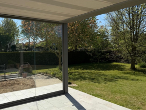 Sous pergola grise store plissé, paroi verre coulissante et dalles face au jardin