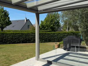 Pergola alu grise toit verre, terrasse carrelée, haie et toit gris en arrière-plan