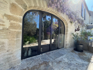 Baie cintrée anthracite quatre vantaux dans arche pierre, glycine violette au-dessus