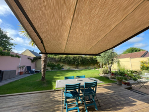 Store banne beige déployé sur terrasse bois, table bleue et palmiers au jardin