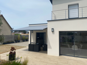 Store banne gris sur terrasse carrelée beige, salon rotin noir sous façade blanche