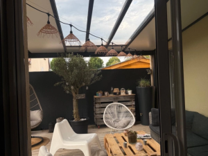 Pergola anthracite toit vitré, store vertical crème, guirlandes et salon palettes sur deck