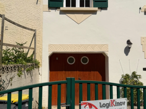 Porte bois lames à hublots ronds, linteau pierre sculpté, volets verts derrière grillage