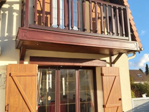 Balcon bois foncé à balustres, volets chêne ouverts et linteau bois sous crépi