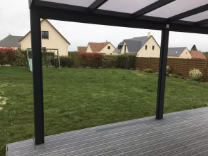 Sous pergola anthracite, terrasse composite grise, pelouse et maisons voisines par temps gris