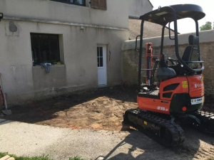 Mini-pelle orange sur sol marque rouge, devant facade beige en terrassement