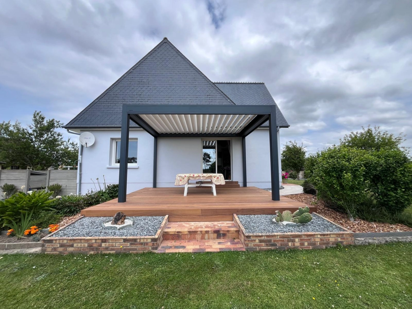 Pergola bioclimatique anthracite, lames blanches, terrasse bois surélevée devant maison blanche