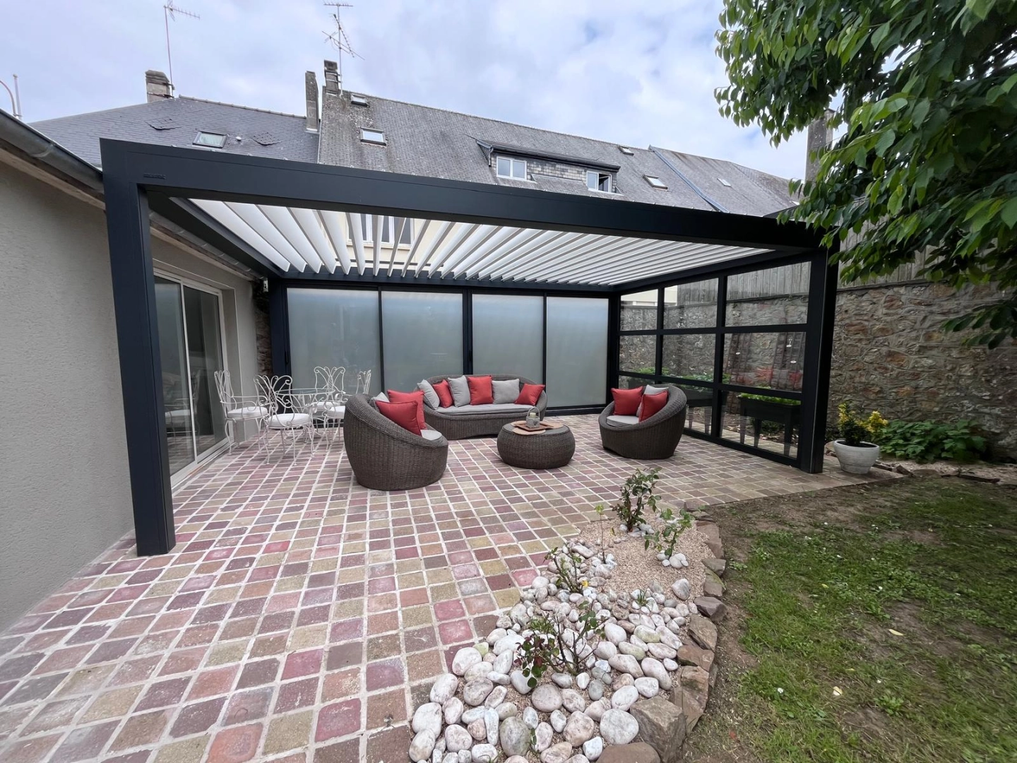 Pergola bioclimatique noire, lames blanches, parois verre dépoli et salon rotin sur pavés