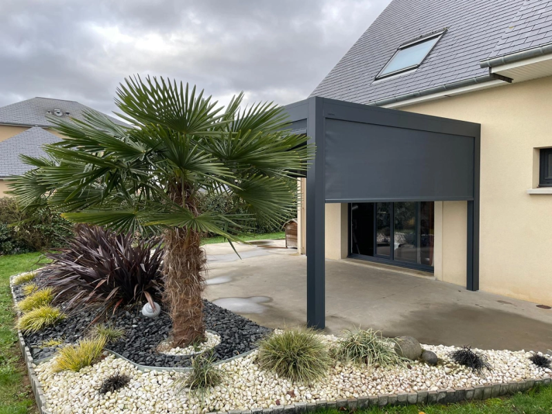 Pergola adossée anthracite, store latéral gris, dalle béton, massif pierres et palmier