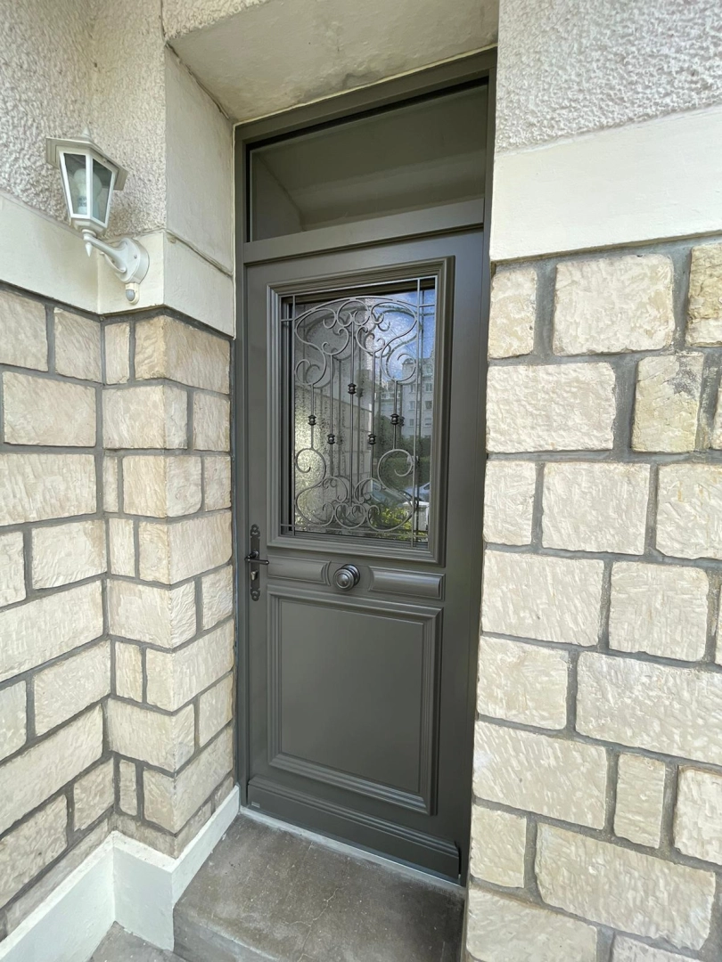 Porte d'entrée anthracite, vitrage avec grille fer forgé, mur pierre beige et imposte