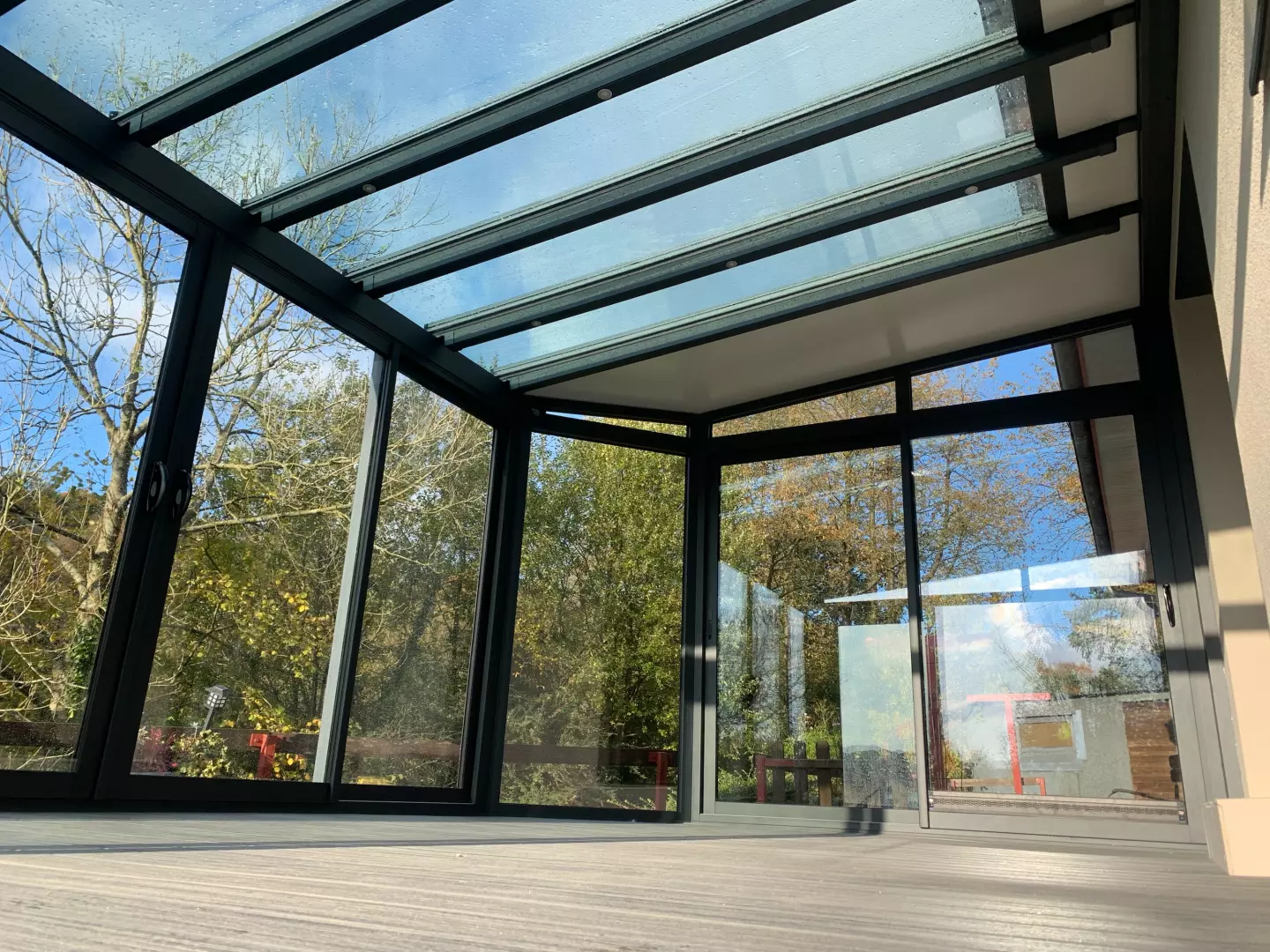 Véranda alu anthracite, parois et toit vitrés, sol en lames grises, vue jardin boisé