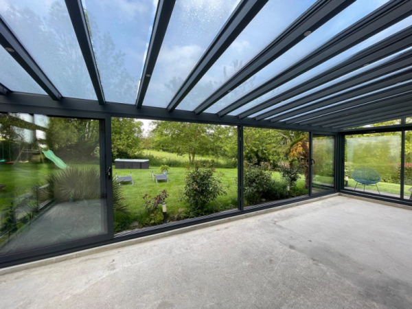 Véranda anthracite : dalle brute, toit verre incliné, vue sur jardin verdoyant