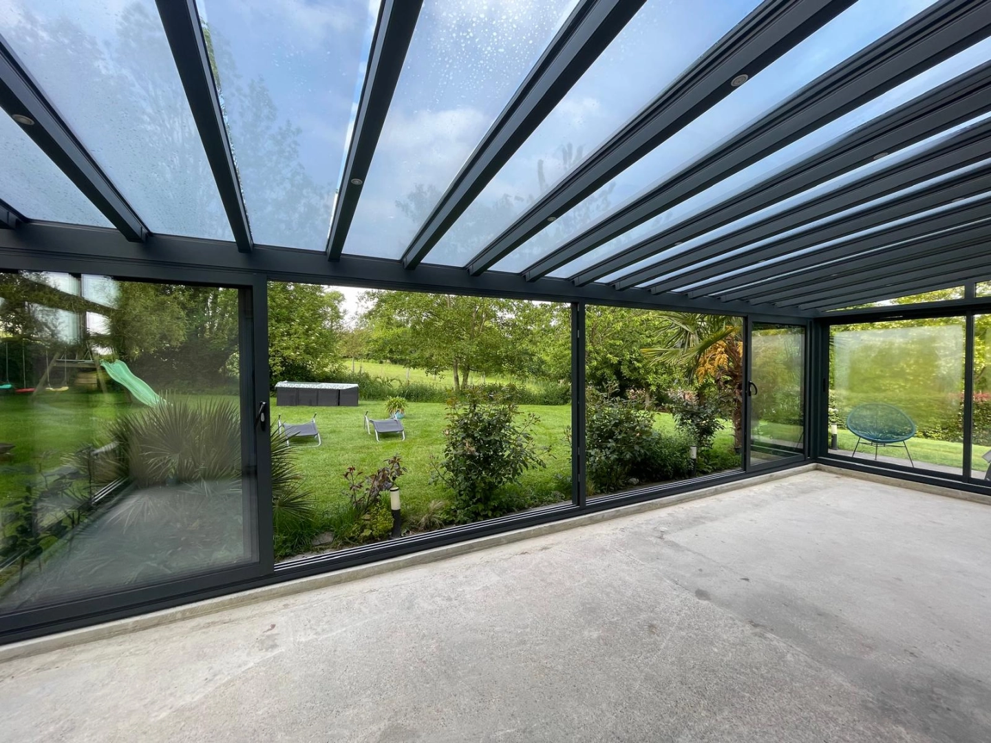 Véranda anthracite : dalle brute, toit verre incliné, vue sur jardin verdoyant