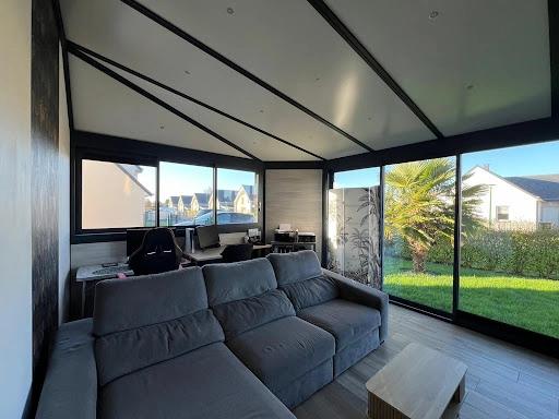 Véranda intérieure alu noir, plafond rayonnant, bureau et canapé gris