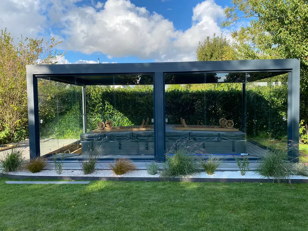 Pergola vitrée alu anthracite autour d'un spa couvert, terrasse bois, pelouse et haie
