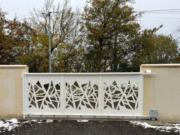 Portail coulissant blanc motifs géométriques ajourés, murets enduits, gravier humide