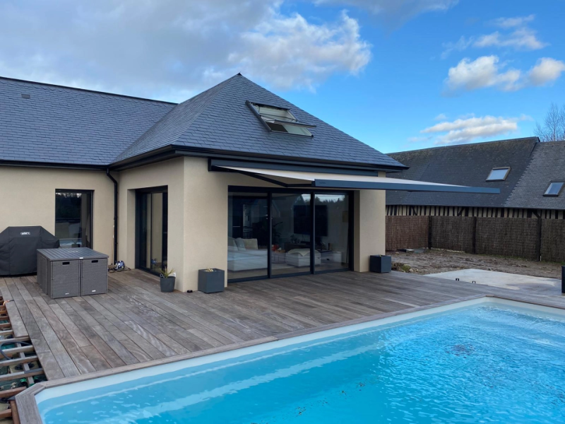 Store banne anthracite déployé sur terrasse bois, piscine bleue et toiture ardoise