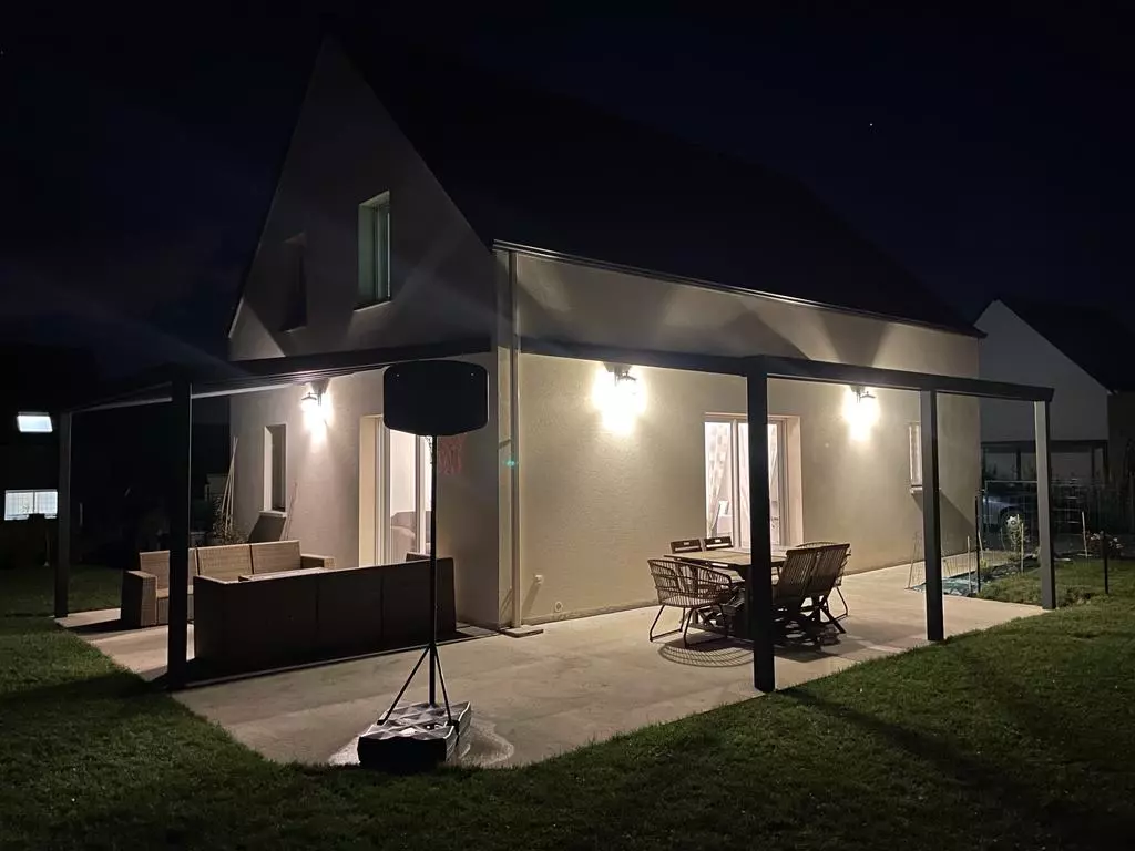 Pergola alu noir toit plat, terrasse dalles claires éclairée, maison crème la nuit