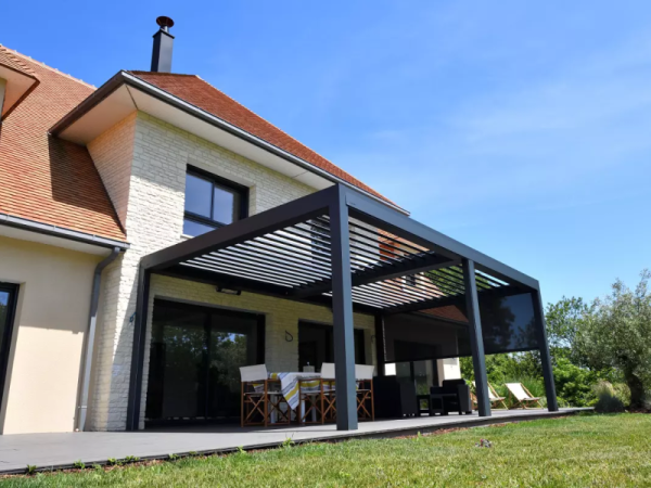 Pergola bioclimatique anthracite, lames orientables et store latéral, terrasse devant maison en pierre