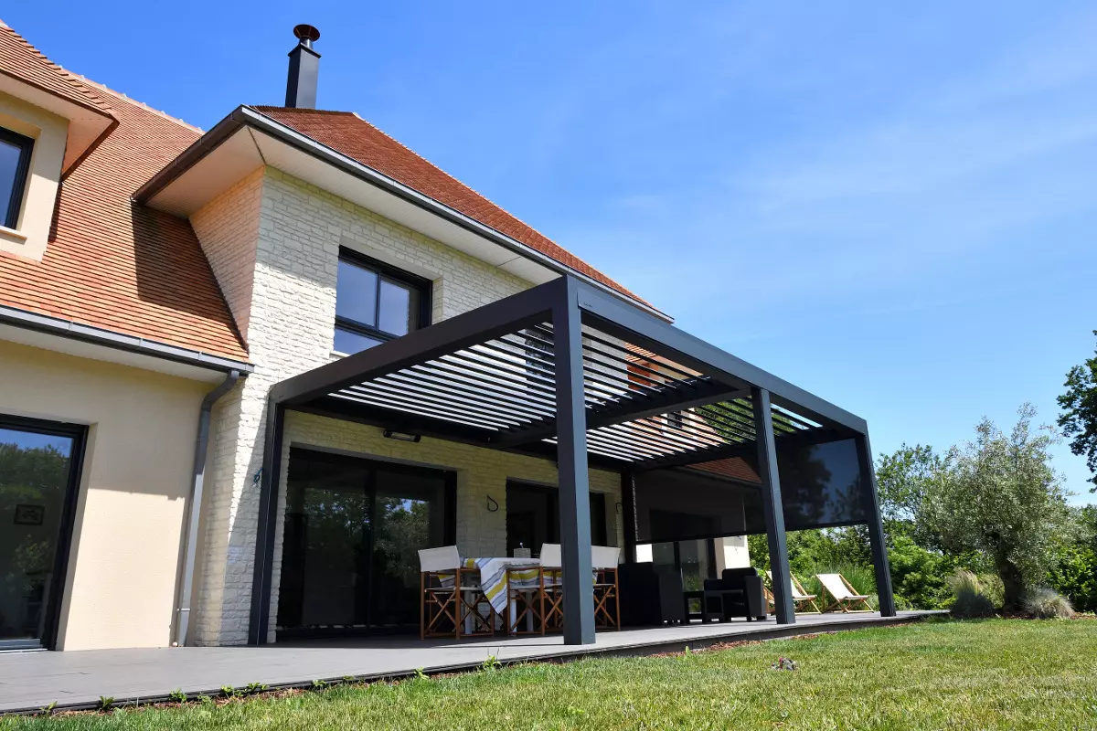 Pergola bioclimatique anthracite, lames orientables et store latéral, terrasse devant maison en pierre