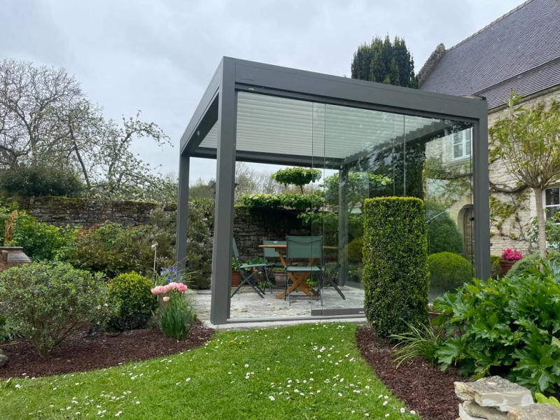 Pergola bioclimatique anthracite, lames claires, parois vitrées, jardin pierre et pelouse